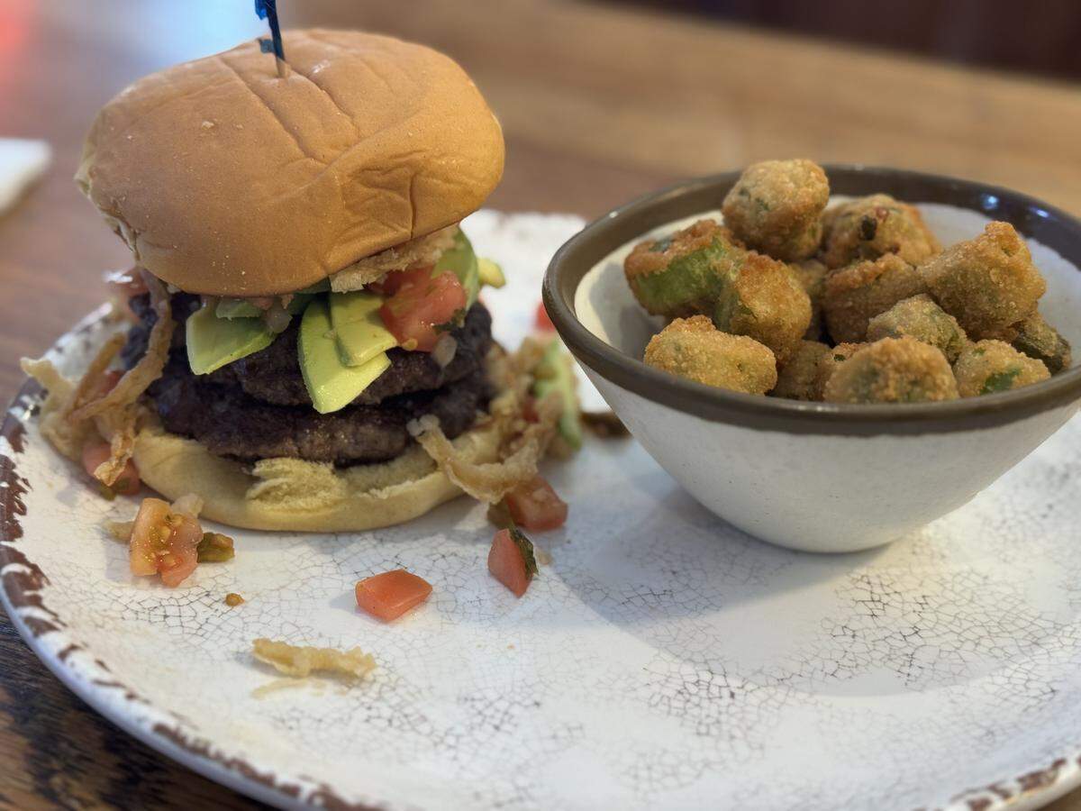 A close-up of a double-patty beef burger topped with thick slices of fresh avocado, diced tomatoes, and crispy fried onions on a toasted bun. A small ceramic bowl filled with golden-brown breaded fried okra sits next to it on a speckled white plate.