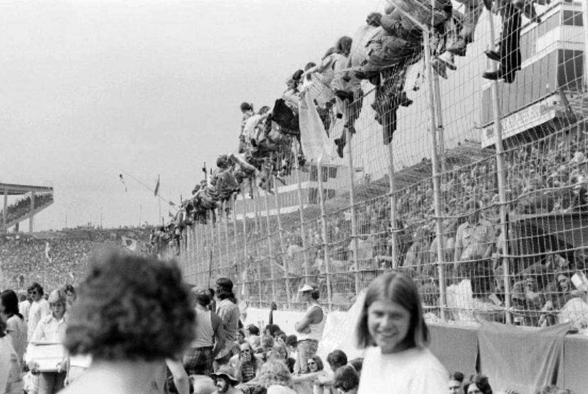 Attendees sitting on top of a fence around the speedway to get a view of the stage.