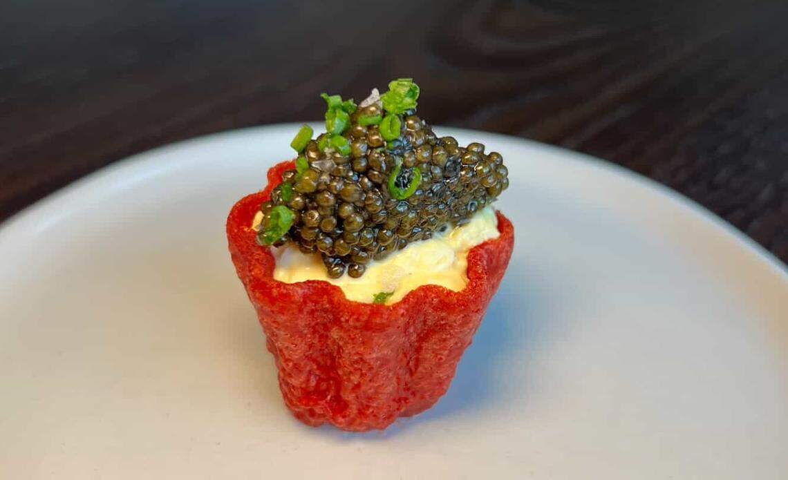 A macro shot of a gourmet amuse-bouche on a white plate. A small, red, cup-shaped crisp with fluted edges holds a savory cream filling. The filling is completely topped with a mound of glossy black sturgeon caviar and a sprinkle of finely chopped chives.