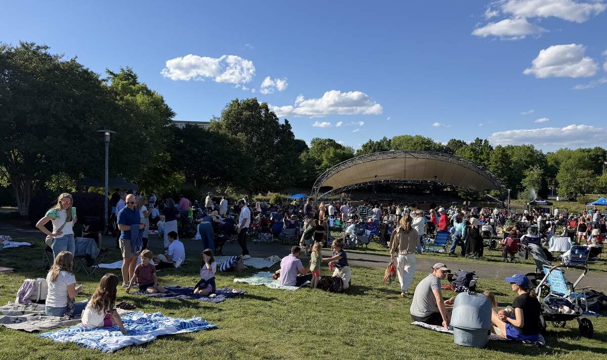 A crowd of people gathered on a grassy lawn with picnic blankets and lawn chairs facing an outdoor amphitheater stage on a sunny day under a blue sky with scattered clouds.