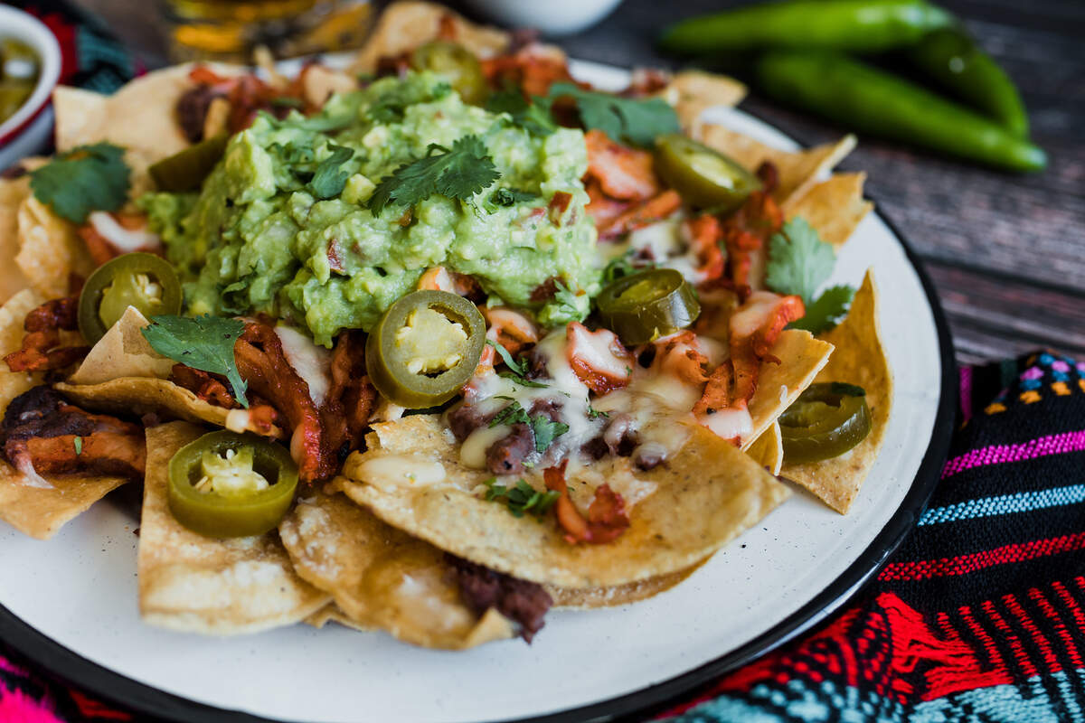 A plate of loaded nachos topped with a large scoop of guacamole, melted white cheese, meat, sliced jalapeños, and fresh cilantro leaves.