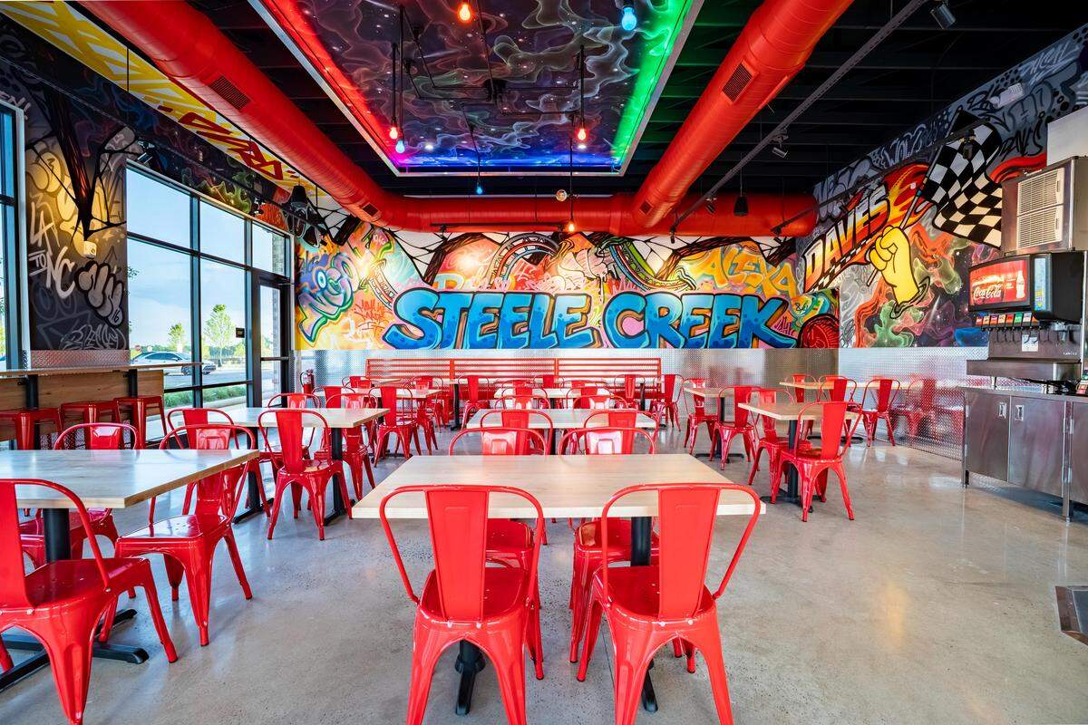 A wide, eye-level indoor shot of a vibrant restaurant dining room filled with rows of light wood tables paired with bright red metal Tolix-style chairs. The space is heavily decorated with colorful, street-art style graffiti murals covering the walls, with the words “STEELE CREEK” painted in large blue bubble letters on the back wall. The ceiling features exposed black ductwork alongside prominent, bright red HVAC pipes and a central recessed ceiling panel painted like a colorful galaxy with neon lighting. Polished concrete flooring runs throughout, and a stainless steel soda fountain station sits on the far right.