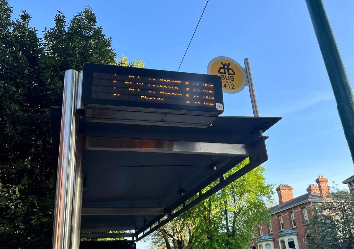 A slightly low-angle shot of a bus stop in Dublin, Ireland. The bus stop has a digital display board showing bus routes and times, though the text is blurred. A yellow sign with a bee logo reads “BUS STOP 412” and “TFI.” The bus stop is made of metal and glass, and in the background, a leafy green tree and the red brick rooftops of residential buildings are visible under a clear blue sky.