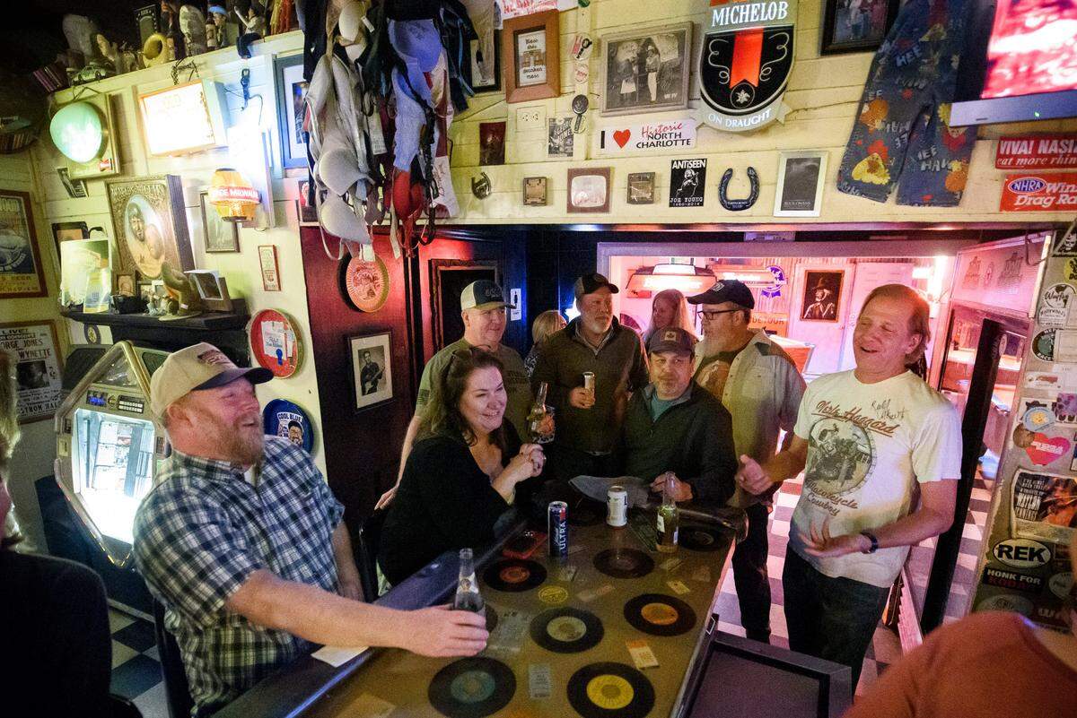 A group of friends gathers around a long table in a dimly lit, eclectic bar. The table has a built-in vintage jukebox or record player, with old 45 rpm records visible on its surface. The bar is decorated with a chaotic collection of memorabilia, including framed pictures, a horseshoe, various signs, and even bras hanging from the ceiling. A man in a plaid shirt sits in the foreground, smiling. Another man in a white t-shirt with a graphic on it stands and gestures while talking. Everyone appears to be enjoying drinks and conversation in the cluttered but cozy atmosphere.