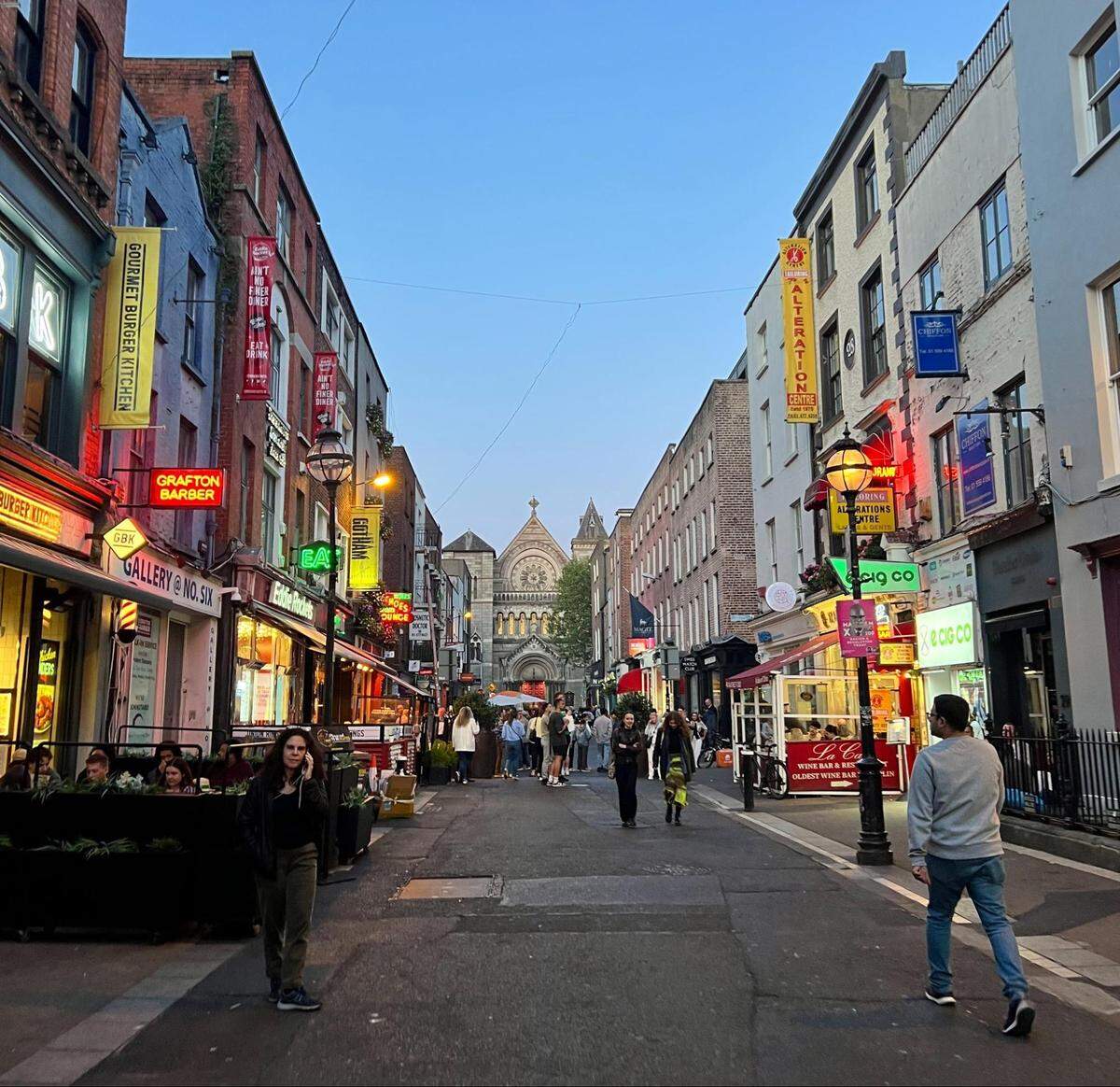 A street view in Dublin, Ireland, in the early evening. The cobblestone street is lined with numerous shops and restaurants with colorful signs, many of which are neon and illuminated. A crowd of people is walking down the street towards a church building in the distance. The sky is a light blue, and the buildings on both sides are tall and narrow.