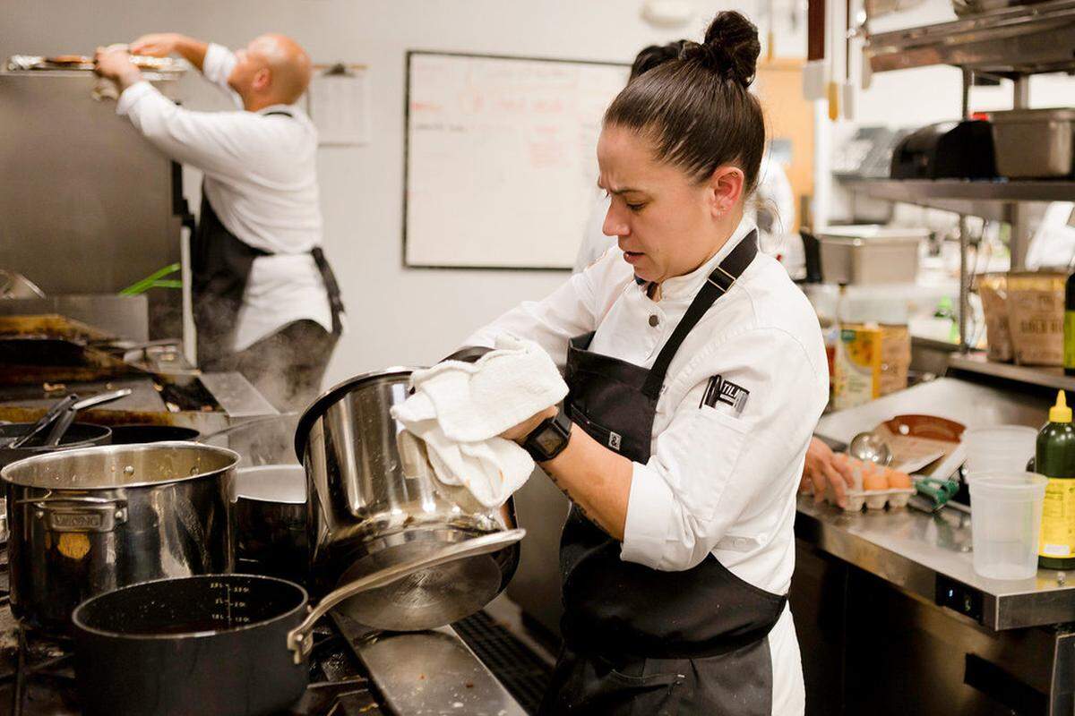 A candid, eye-level shot inside a professional kitchen. In the center, a chef with hair pulled back in a high bun wears a white chef’s coat and a black apron and focuses on wiping or holding a large stainless steel pot with a white towel. In the blurred background, another chef is working near a grill, and various kitchen tools, stainless steel counters, and a white board are visible, creating a busy culinary atmosphere.