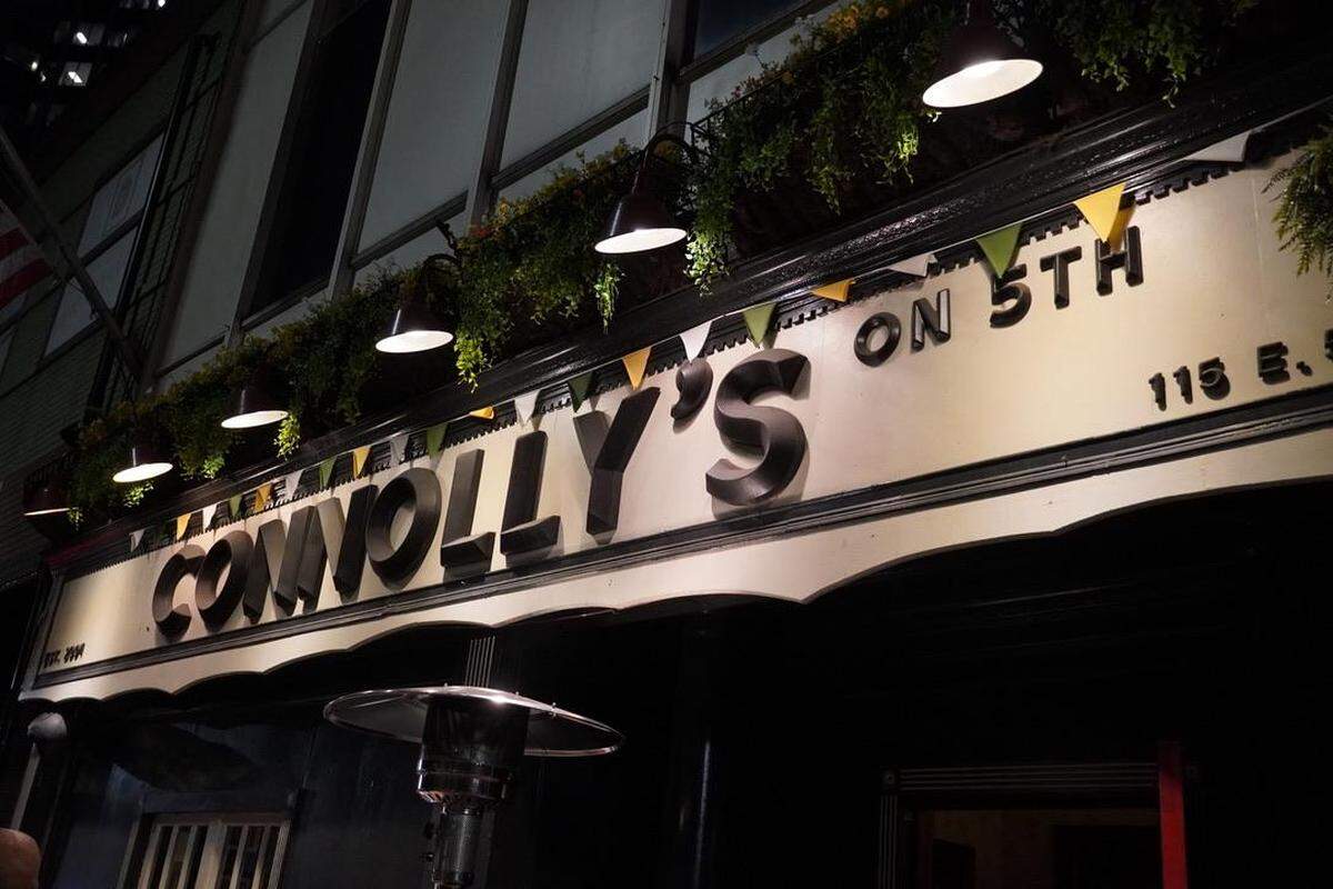 A low-angle, nighttime shot of the illuminated “CONNOLLY’S ON 5TH” pub sign. The sign features large dark letters, is strung with small pennant flags, and is illuminated by a row of black lamps set beneath a planter box filled with greenery.