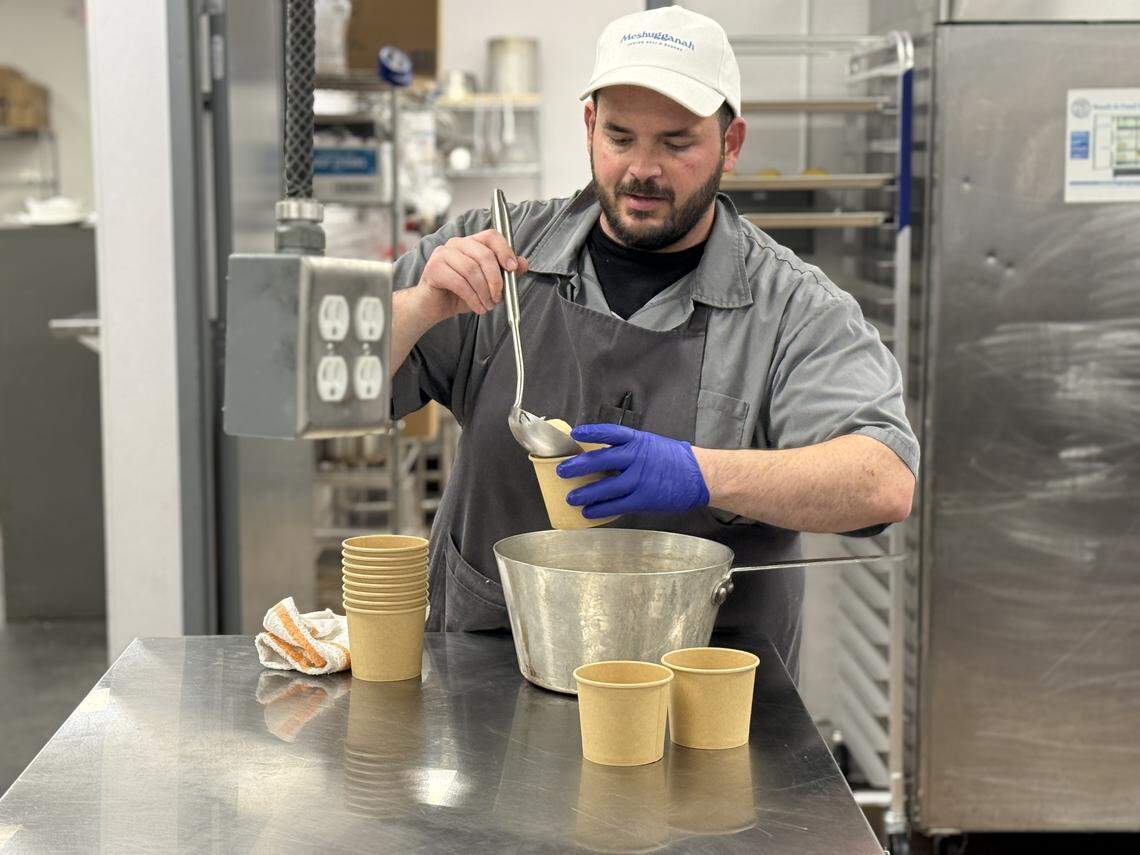 A chef wearing a white “Meshugganah” baseball cap, a grey shirt, and a dark apron carefully ladles a matzo ball from a large metal pot into a small brown paper cup while wearing blue latex gloves and working in a professional kitchen environment.
