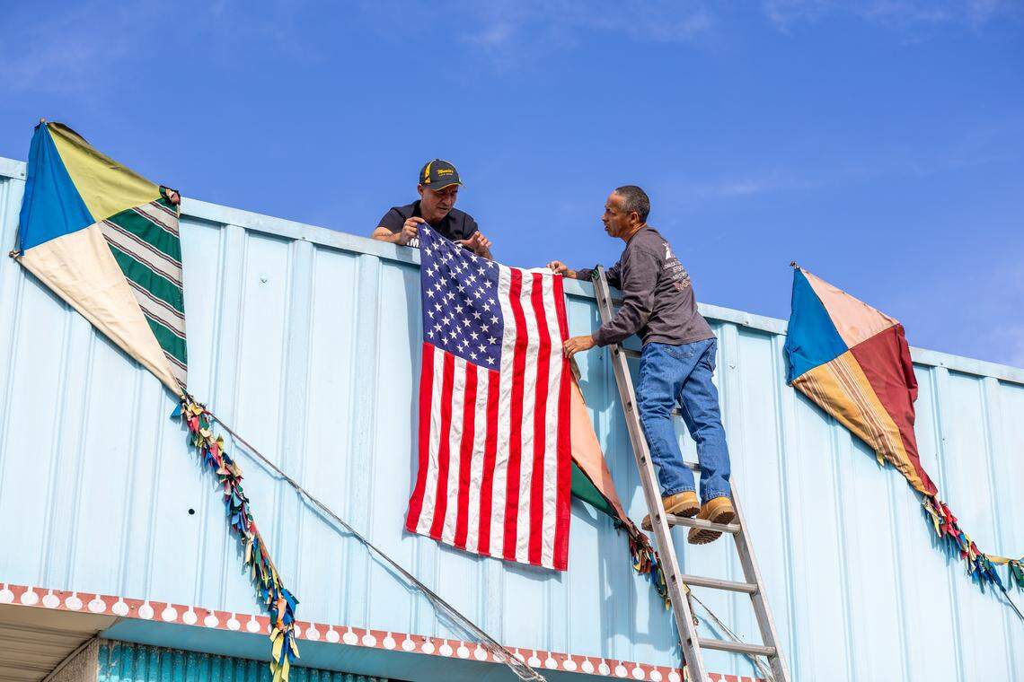 Two men on a ladder affix a large American flag to the blue facade of a Latino-owned bakery in Charlotte. The business owner stands on the roof as the flag is hung. The bakery’s main window is covered in colorful messages and the word “OPEN” is visible on an unlit sign.