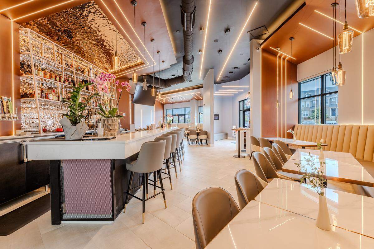 The modern interior of Flame Asian Bistro & Bar featuring a long white marble-topped bar with grey upholstered barstools on the right. The space is decorated with linear LED light strips on the ceiling and walls, several hanging Edison-style pendant lights, and a backlit sign displaying a kanji character. In the background, there is a dining area with tan booth seating and small tables.