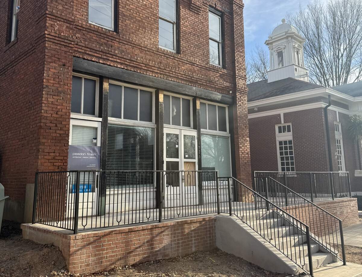 An exterior view of the two-story red brick Leeper & Wyatt building after its relocation. It features a new brick staircase and landing with a black metal railing. A sign for “peadon finein architecture | interiors | planning” is attached to the lower-left window. In the background, a neighboring brick building with a white decorative cupola is visible.