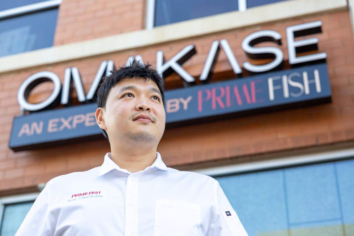 A low-angle portrait of a chef standing outside in front of a brick building. Above the chef is a prominent sign that reads “OMAKASE BY PRIME FISH.”