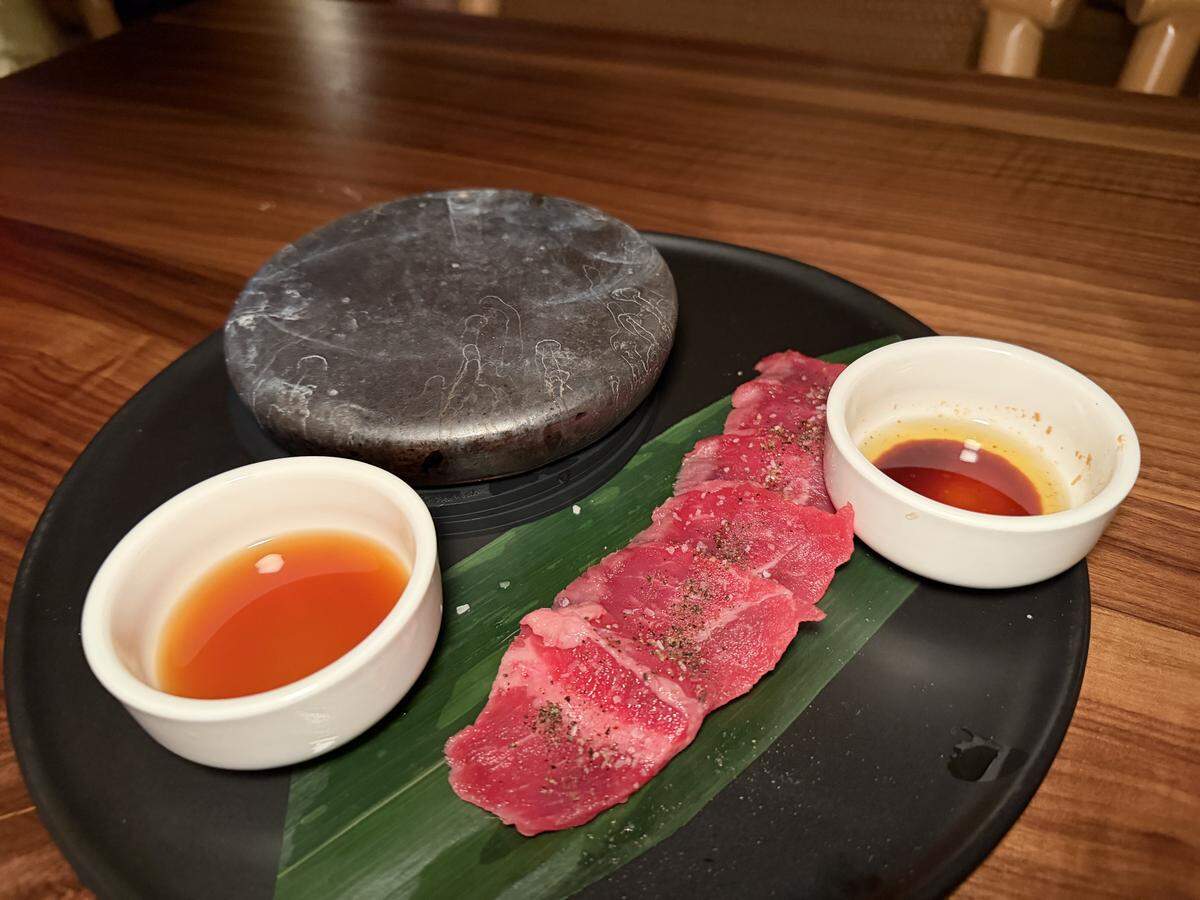An interactive DIY dining course, likely Ishi-yaki (stone-grilled) wagyu beef, is presented on a matte black plate. The meat is vibrant red and fanned out across a bright green banana leaf seasoned with cracked black pepper and a pinch of sea salt. A thick, weathered hot volcanic stone (or cast-iron puck) sits on the left side of the plate, used for searing the beef to the diner’s preference. Two small white ramekins flank the stone and meat.