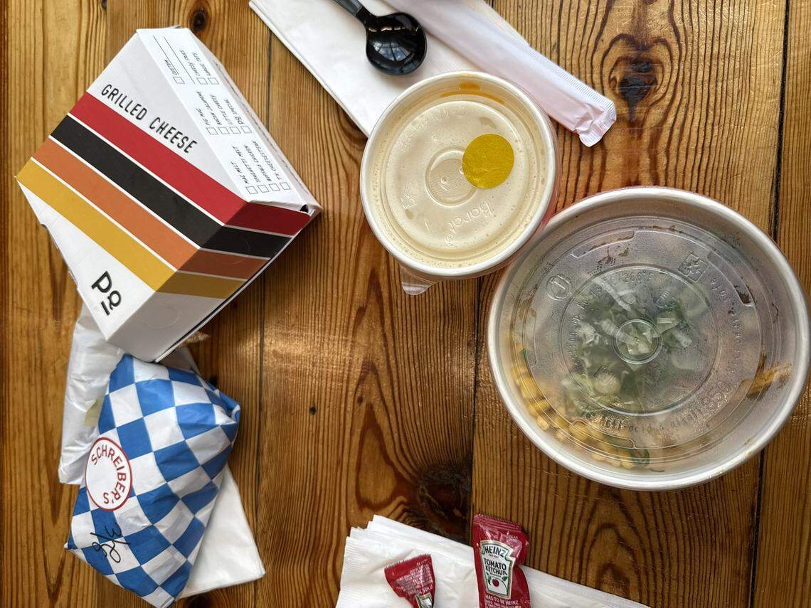 A top-down view of a spread of takeout containers on a light-colored wooden table. On the left is a white box labeled “GRILLED CHEESE” from Papi Queso, featuring a retro orange and brown striped design. Below it is a small sandwich wrapped in blue and white checkered paper with a “Schreiber’s” sticker. To the right are two round plastic containers with translucent lids: one smaller container holding a light-colored beverage or soup, and a larger bowl containing a salad or grain bowl with visible corn and greens. A black plastic spoon and several white napkins are scattered around the food.