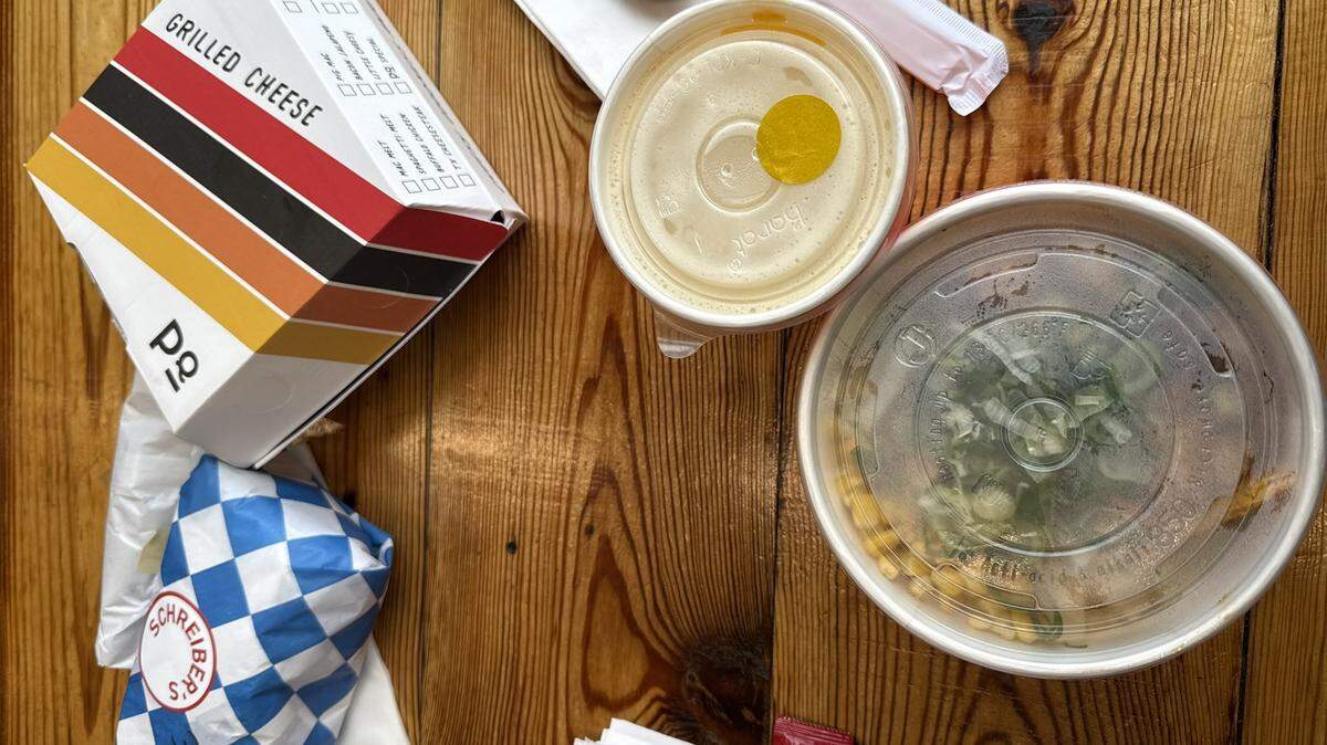 A top-down view of a spread of takeout containers on a light-colored wooden table. On the left is a white box labeled “GRILLED CHEESE” from Papi Queso, featuring a retro orange and brown striped design. Below it is a small sandwich wrapped in blue and white checkered paper with a “Schreiber’s” sticker. To the right are two round plastic containers with translucent lids: one smaller container holding a light-colored beverage or soup, and a larger bowl containing a salad or grain bowl with visible corn and greens. A black plastic spoon and several white napkins are scattered around the food.