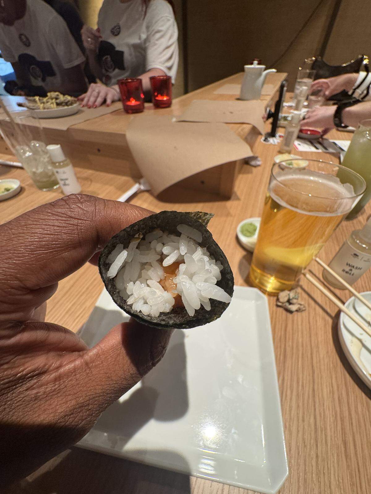 A close-up, first-person view of a hand holding a cylindrical sushi hand roll (temaki). The roll is wrapped in crisp dark seaweed and filled with white sushi rice and a piece of salmon in the center. The background shows a lively wooden sushi bar with a tall glass of beer, chopsticks, and other diners.