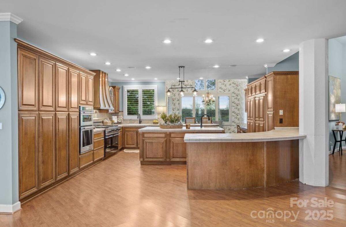 A wide-angle interior view of a spacious, light-filled kitchen featuring extensive, floor-to-ceiling medium-brown wood cabinetry, wood floors, and light blue walls. The kitchen includes a central island with a white countertop and a breakfast bar extending from it. Appliances are stainless steel, and a window with white shutters is located above the main sink area.