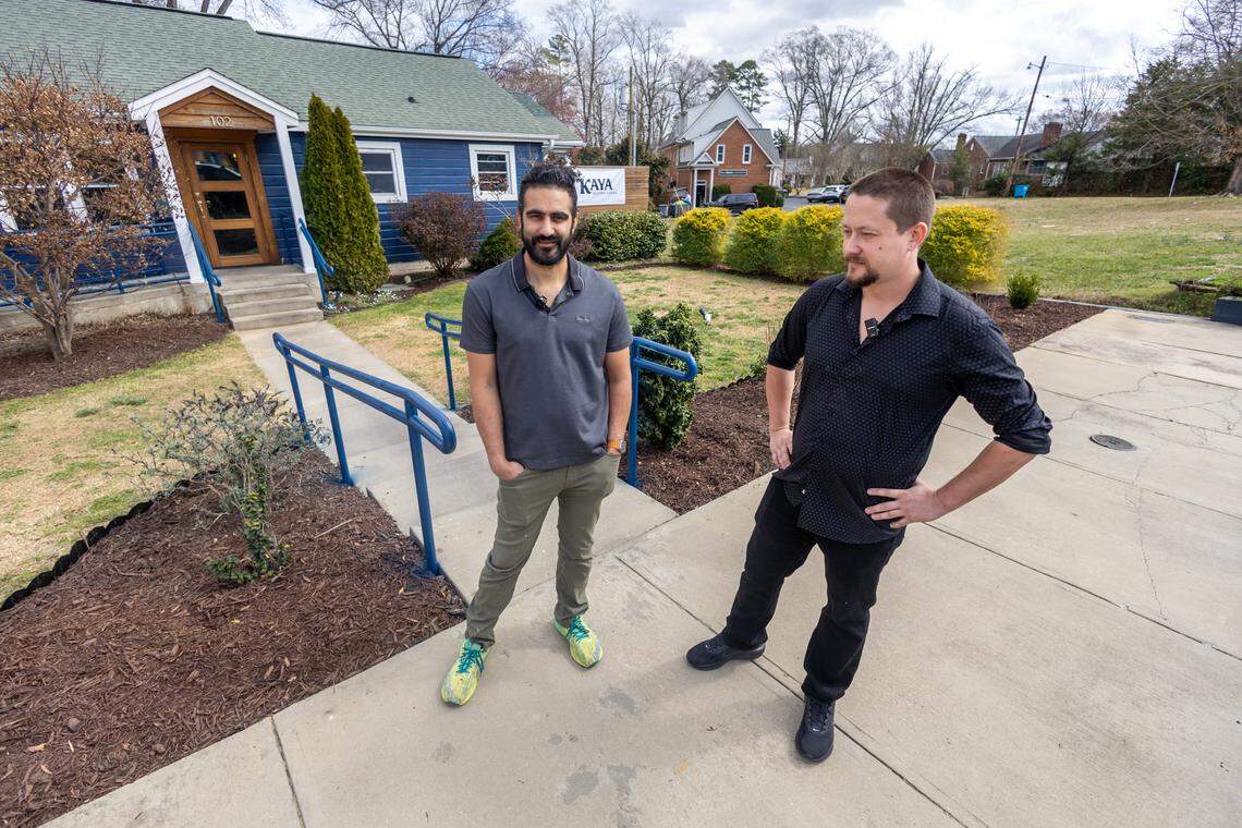 Two restaurateurs are standing and talking on a concrete sidewalk outside a small, blue building with a sign that reads “KAYA.” The person on the left has a beard and is wearing a gray polo shirt, green pants, and bright yellow-green sneakers. The individual on the right, with a beard and tattoos on his arms, is wearing a black patterned shirt and black pants, with his hand on his hip. A blue railing runs along a ramp leading to the building’s front door. The ground around the building is covered with brown mulch and small bushes. The sky is overcast and the trees are bare.