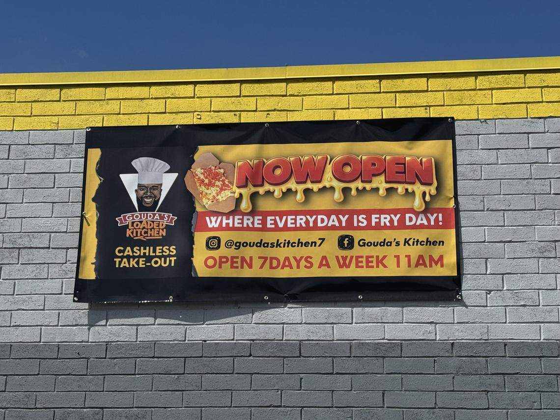 A “Now Open” banner for “Gouda’s Loaded Kitchen” hanging on a grey and yellow brick wall. The sign features a chef logo and the slogan “Where everyday is Fry Day!” along with social media handles and hours.