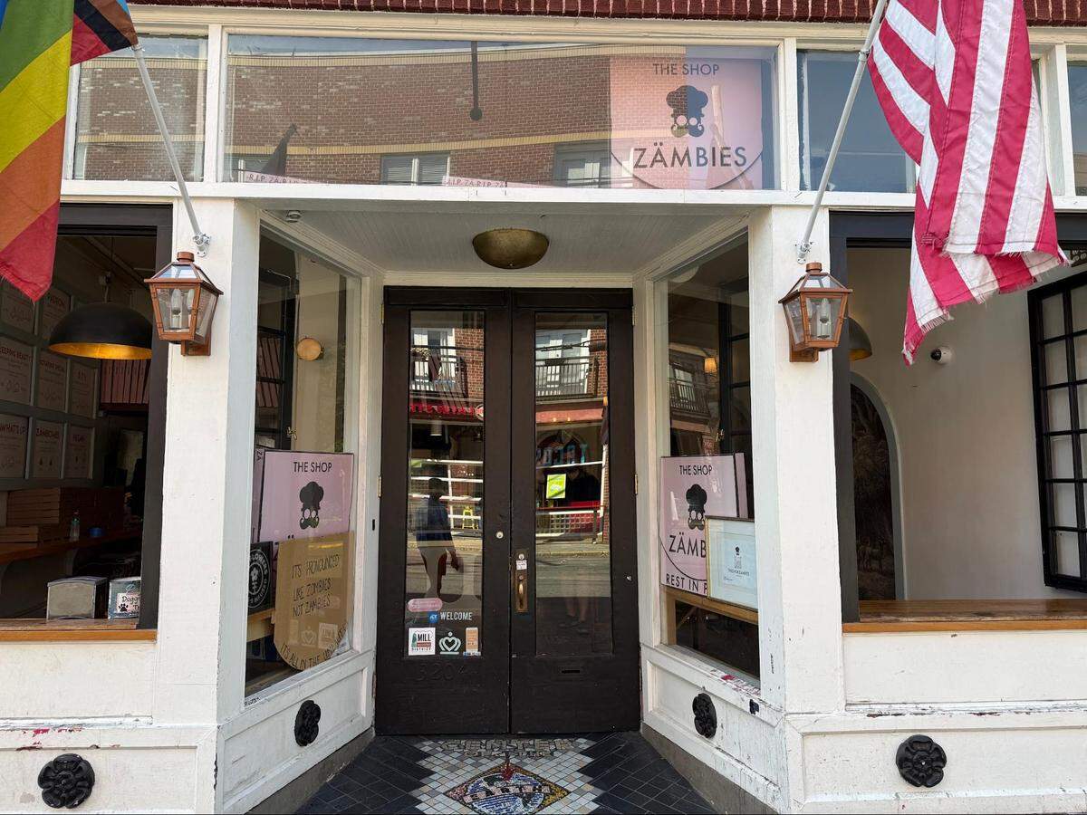 A straight-on exterior shot of the entrance to a pizzeria named “The Shop Zämbies.” The storefront features a white wooden facade with large glass display windows flanking a set of dark double doors. Pink signs with the restaurant’s skull-and-chef-hat logo are visible inside the windows, alongside a hand-drawn cardboard sign clarifying the name’s pronunciation. A pride flag hangs on the left side of the entrance, an American flag hangs on the right, and a decorative mosaic tile design is embedded in the ground right before the doorway.