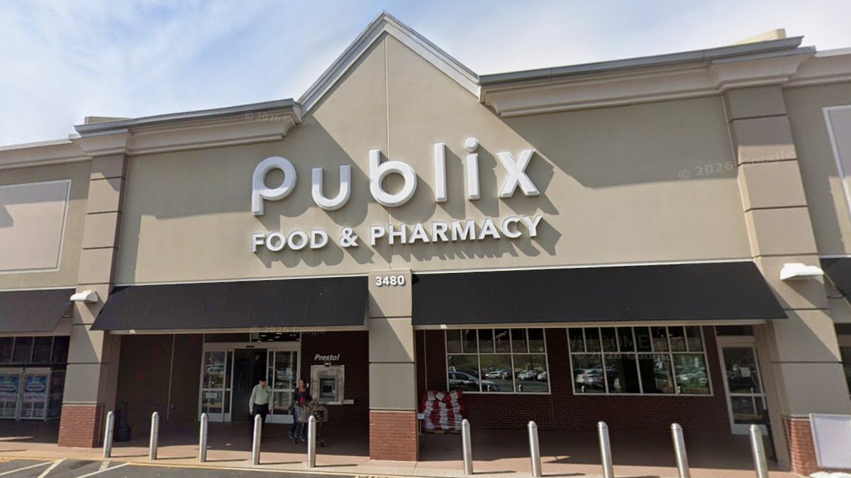 The exterior storefront of a Publix Food & Pharmacy is shown, featuring a beige facade with large white lettering and a black awning. The street address “3480” is visible above the entrance, where a few people are walking near a Presto! ATM. To the left, several silver bollards line the sidewalk in front of the building under a clear blue sky.