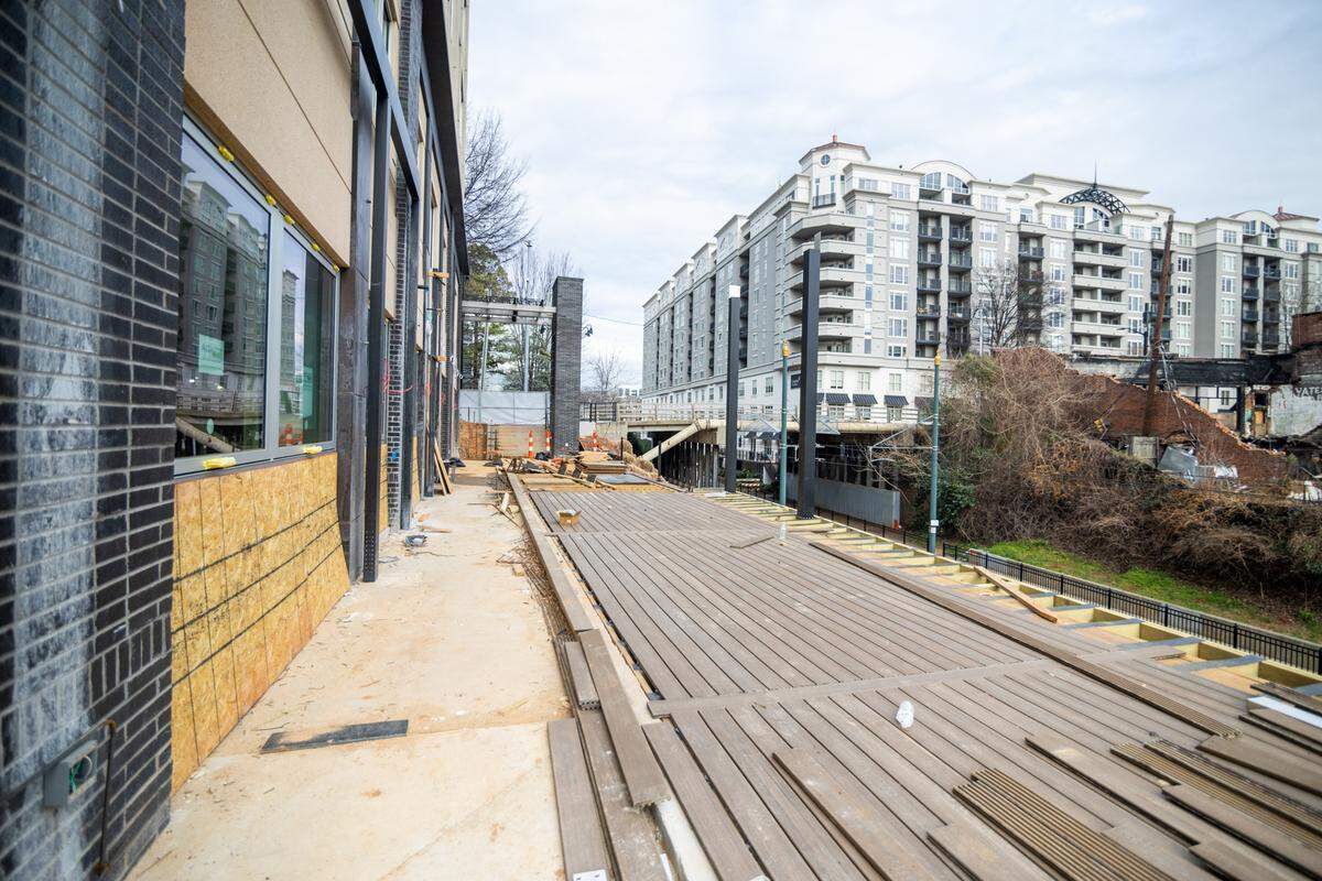 A wide-angle outdoor shot of a construction site showing a long, elevated wooden deck under assembly next to a modern building. To the right, a view of the city includes a multi-story apartment complex and a bridge over light rail tracks.