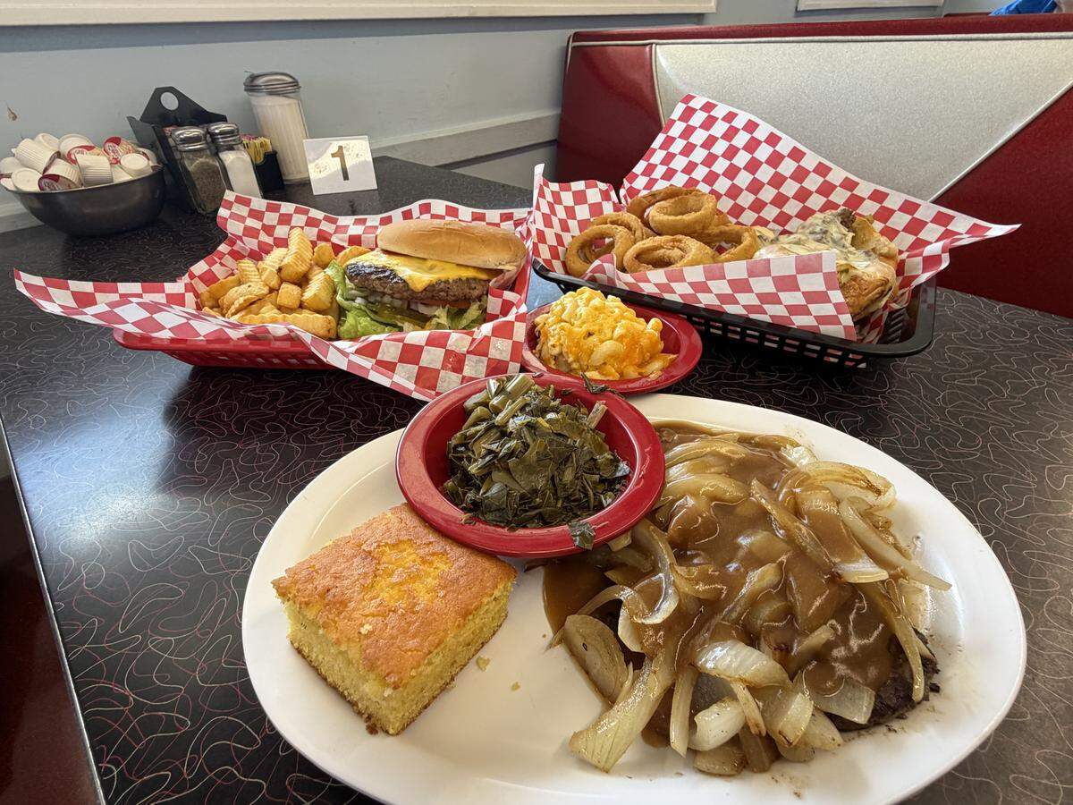 A wide shot of a dining table at Shuffletown Grill. In the foreground is a white plate holding a hamburger steak covered in brown gravy and sautéed onions, served with a thick slice of yellow cornbread and small red bowls of collard greens and macaroni and cheese. In the background, baskets with a cheeseburger and a Philly cheesesteak are visible next to a classic red and silver diner booth.
