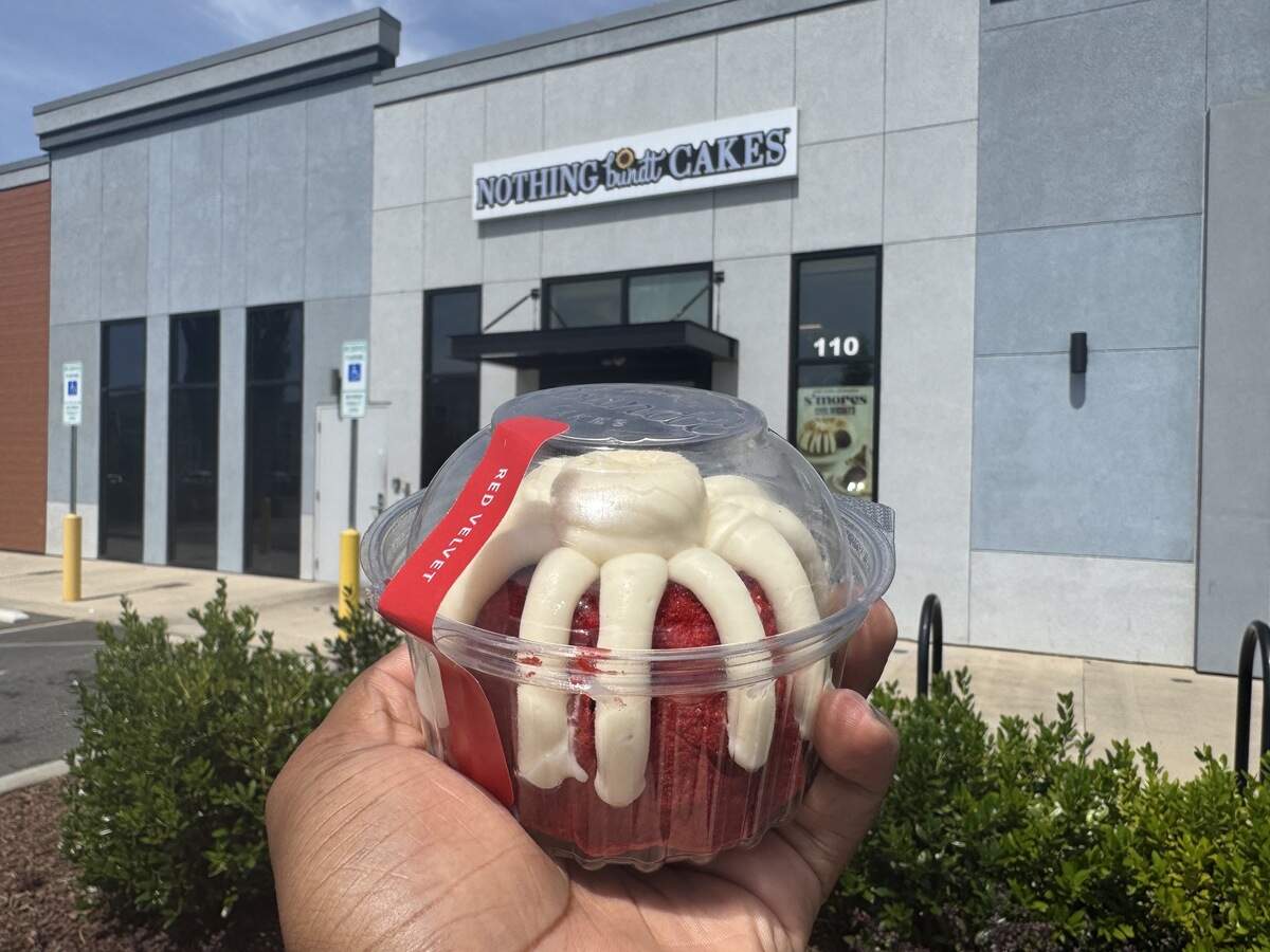 A hand holds a packaged individual-sized red velvet bundt cake with thick white cream cheese frosting from Nothing Bundt Cakes. The actual Nothing Bundt Cakes storefront and parking lot are visible in the bright daylight behind it.