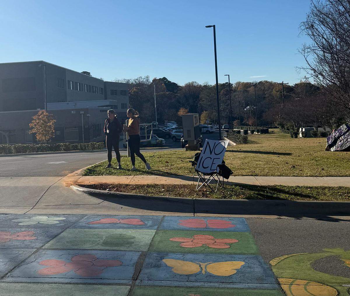 Laura Blum and Patricia Hoke stand outside of Shamrock Gardens Elementary School Monday with a sign in a chair saying “NO ICE.” They are among parents participating in a volunteer effort to carpool kids and stand watch near schools.
