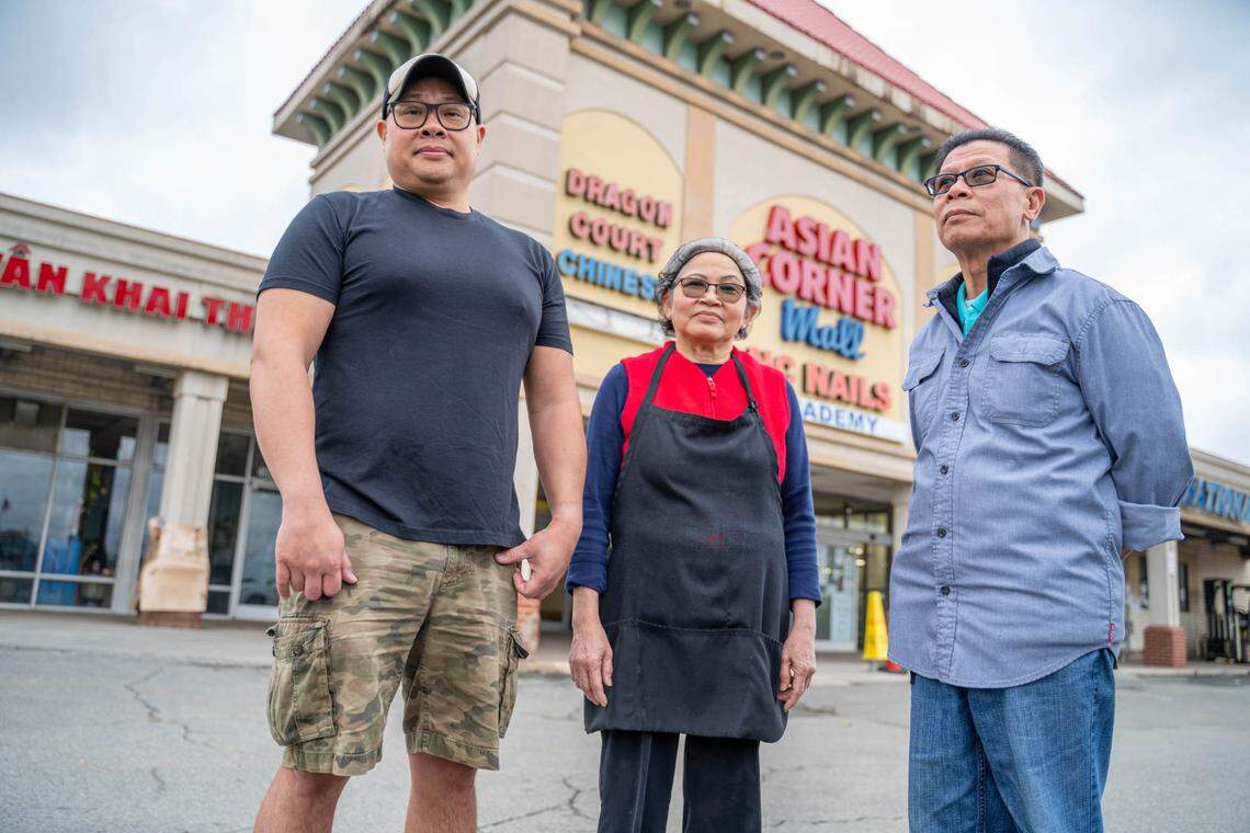 Three people stand for a low-angle portrait in the parking lot of the Asian Corner Mall, a large, beige stucco building with a prominent sign in red and blue letters. The person on the left wears a backward cap, the person in the center wears a black apron, and the person on the right looks away from the camera.