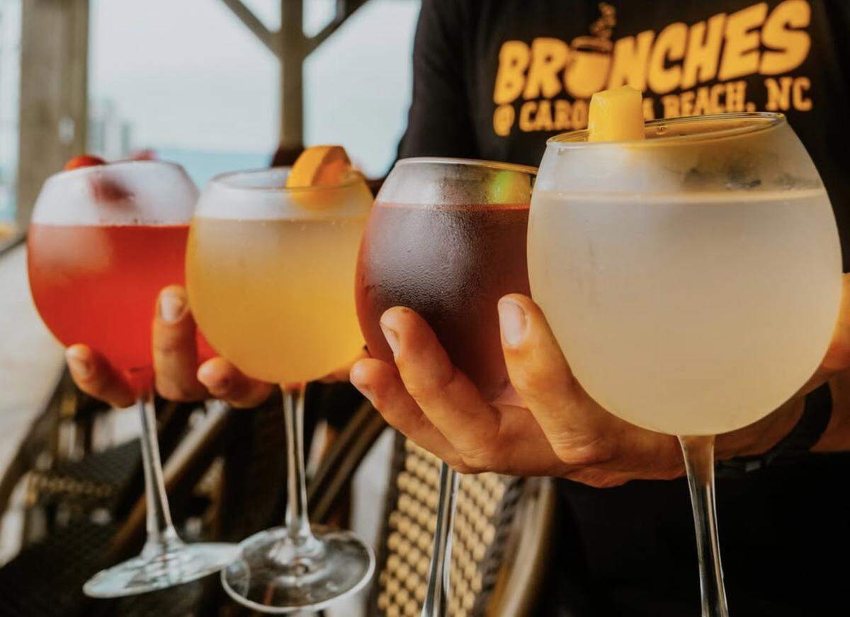 A close-up shot of a person holding four large, round wine glasses filled with colorful brunch cocktails. From left to right, the chilled drinks include a bright red cocktail topped with berries, a vibrant yellow-orange mimosa garnished with a citrus wedge, a deep reddish-purple sangria, and a translucent white cocktail garnished with a piece of pineapple. The person is wearing a black T-shirt featuring the yellow logo for “Brunches @ Carolina Beach, NC.”