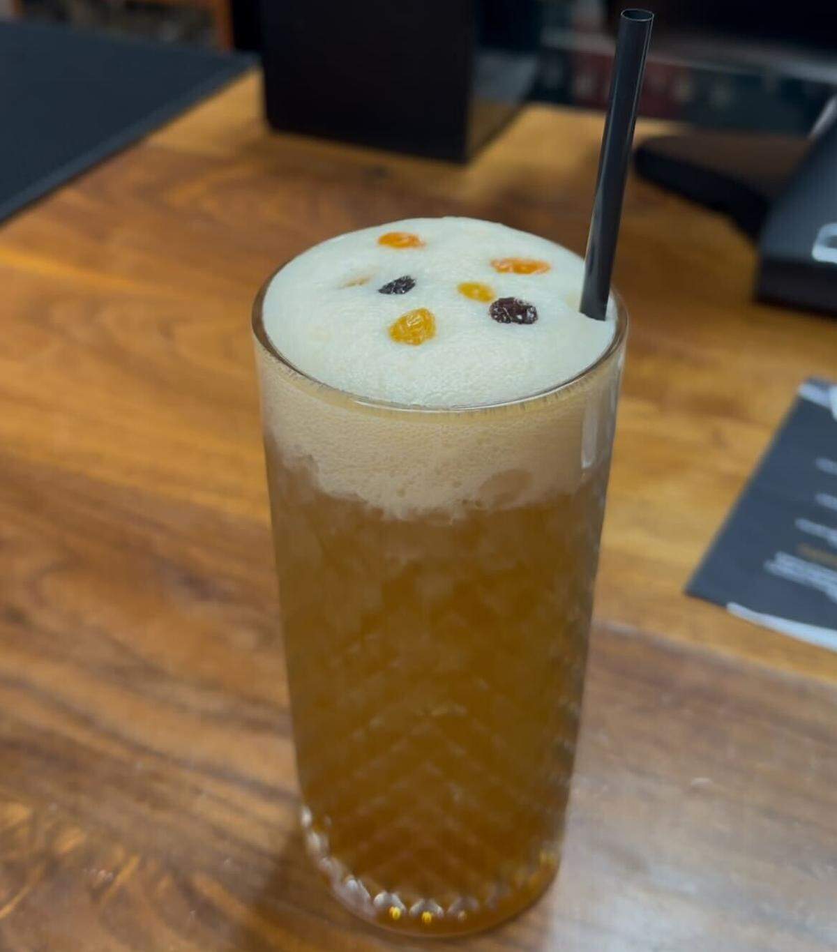 A sophisticated cocktail sits on a wooden table. The tall, patterned glass is filled with an amber-colored liquid and topped with a luxurious, thick cap of white foam. Floating delicately on the foam are a few raisins and vibrant droplets of orange and red bitters.