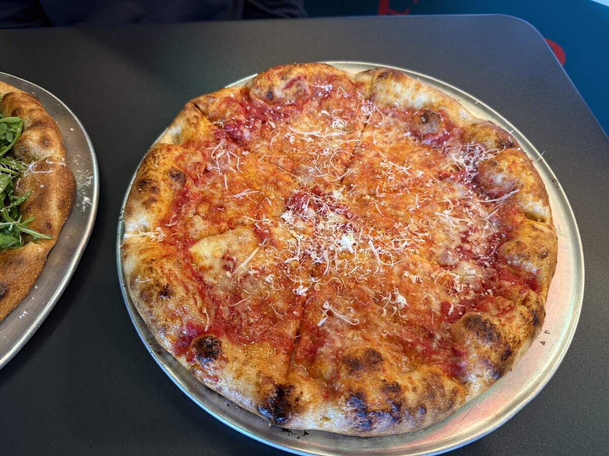 A close-up view of a whole, uncut cheese pizza served on a round metal tray resting on a dark tabletop. The pizza has a thick, puffy, deeply browned crust with charred blisters. The center features rich red tomato sauce and a light dusting of finely grated white cheese. The edge of another pizza topped with fresh green arugula is visible on the left side of the frame.