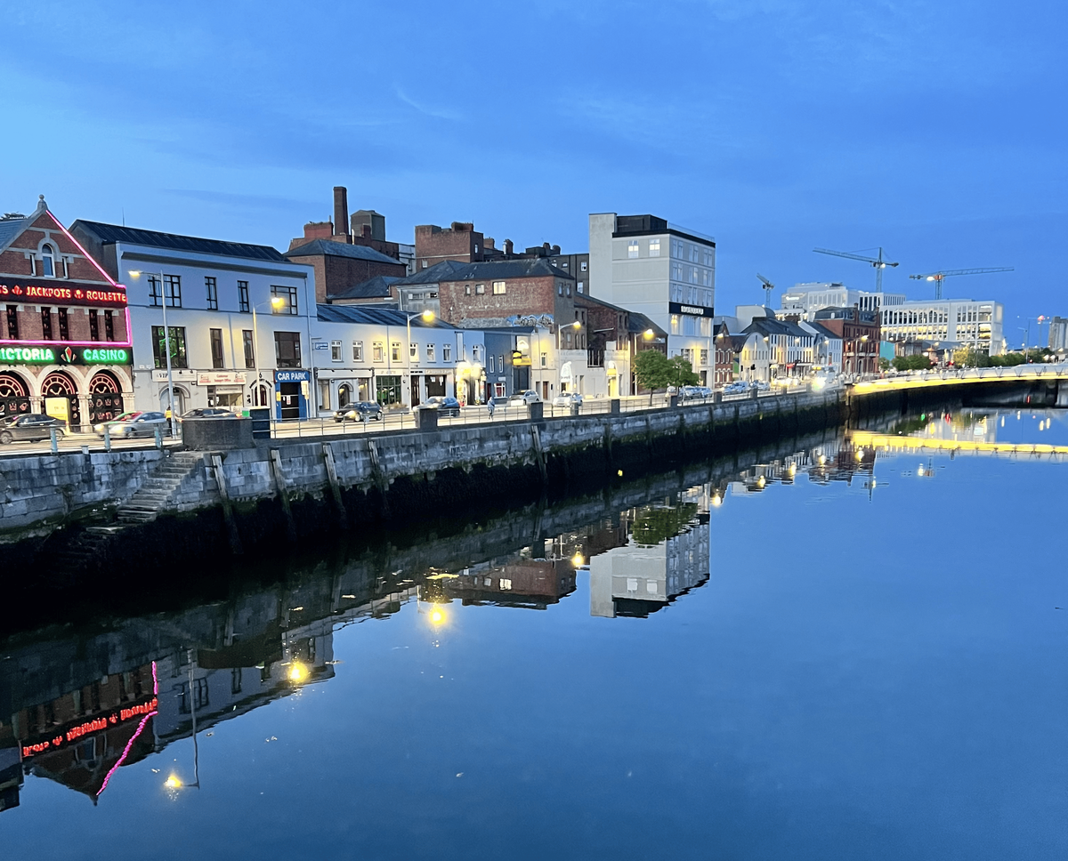 A scenic view of a city waterfront at dusk. A row of buildings of various heights and colors, with lights on, lines the water’s edge. Some of the buildings have neon signs, including one for a casino. The lights from the buildings and a bridge in the distance are reflected in the still, dark water of the harbor. The sky is a deep blue.