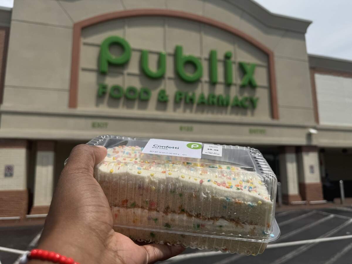 A person’s hand holding a clear plastic container with a Publix bakery confetti bar cake in front of a blurry Publix Food & Pharmacy storefront. The cake features white frosting with colorful rainbow sprinkles on top.