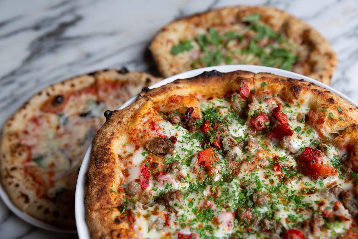 A close-up shot of three round pizzas on a marble countertop. The pizza in the foreground is in focus, topped with melted cheese, sausage, red bell peppers, and fresh herbs. The crust is thick, bubbly, and slightly charred. Two other pizzas, out of focus, are visible in the background: one appears to be a margherita pizza, and the other is topped with green herbs.
