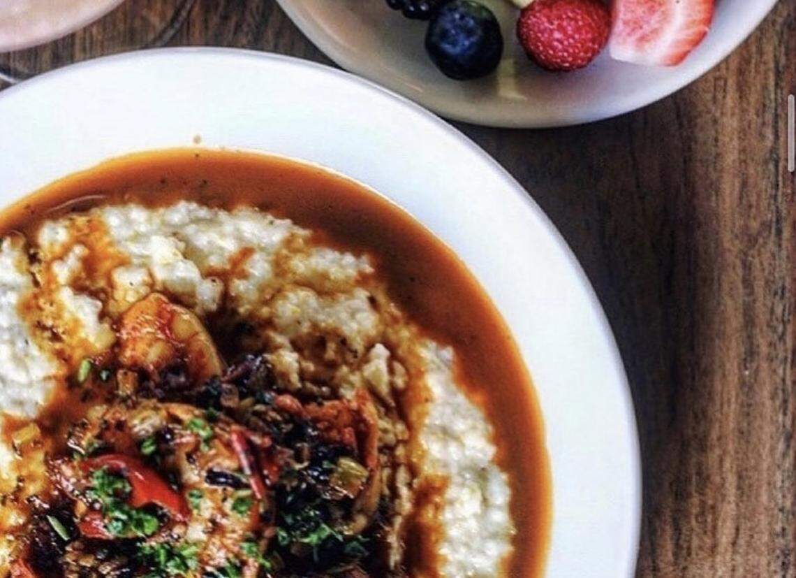 A top-down, close-up shot of a bowl of creamy grits topped with a savory red-sauced shrimp dish and fresh herbs. In the upper portion of the frame, a small side bowl contains fresh blueberries, raspberries, and a strawberry slice.