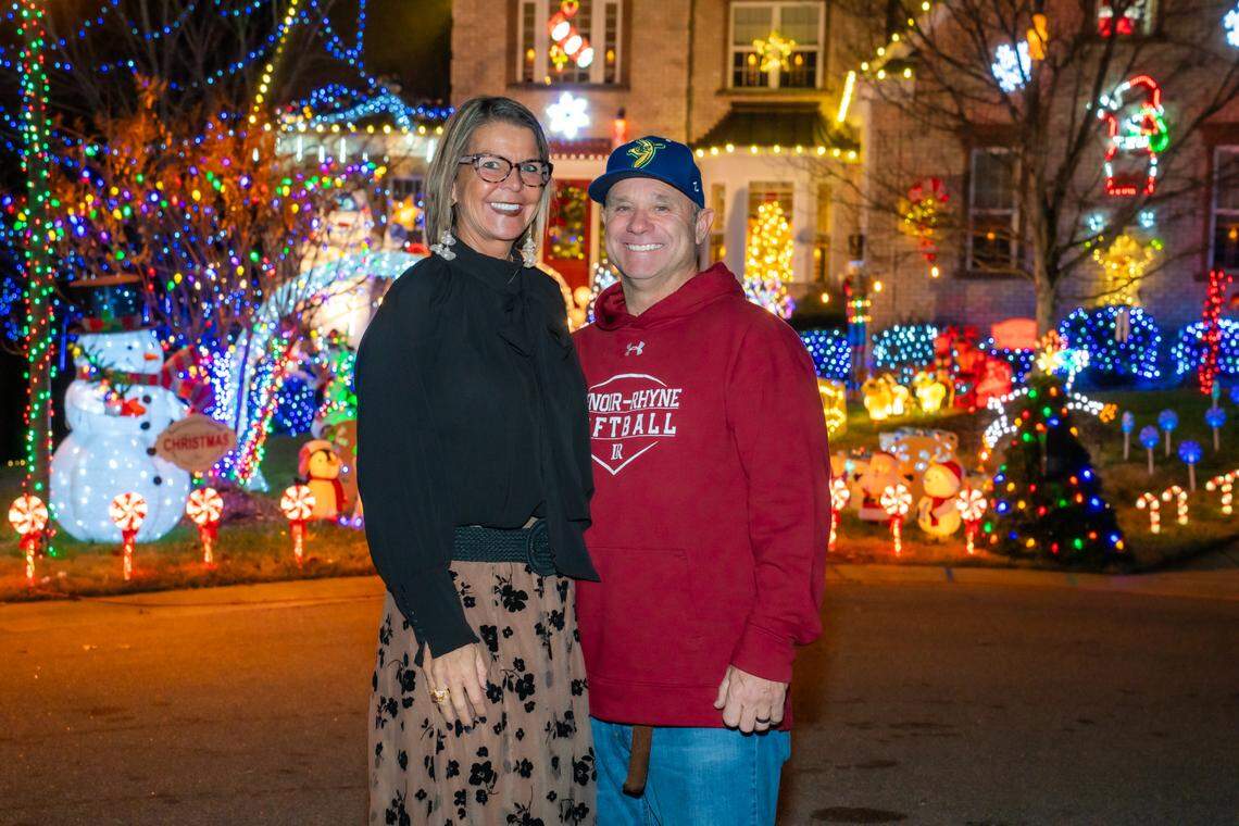 A couple smiling and posing for a photo in front of their home, which is covered in an elaborate and colorful holiday light display. The foreground features numerous small glowing snowmen, candy canes, and blue lights, with the house façade brightly lit in the background.
