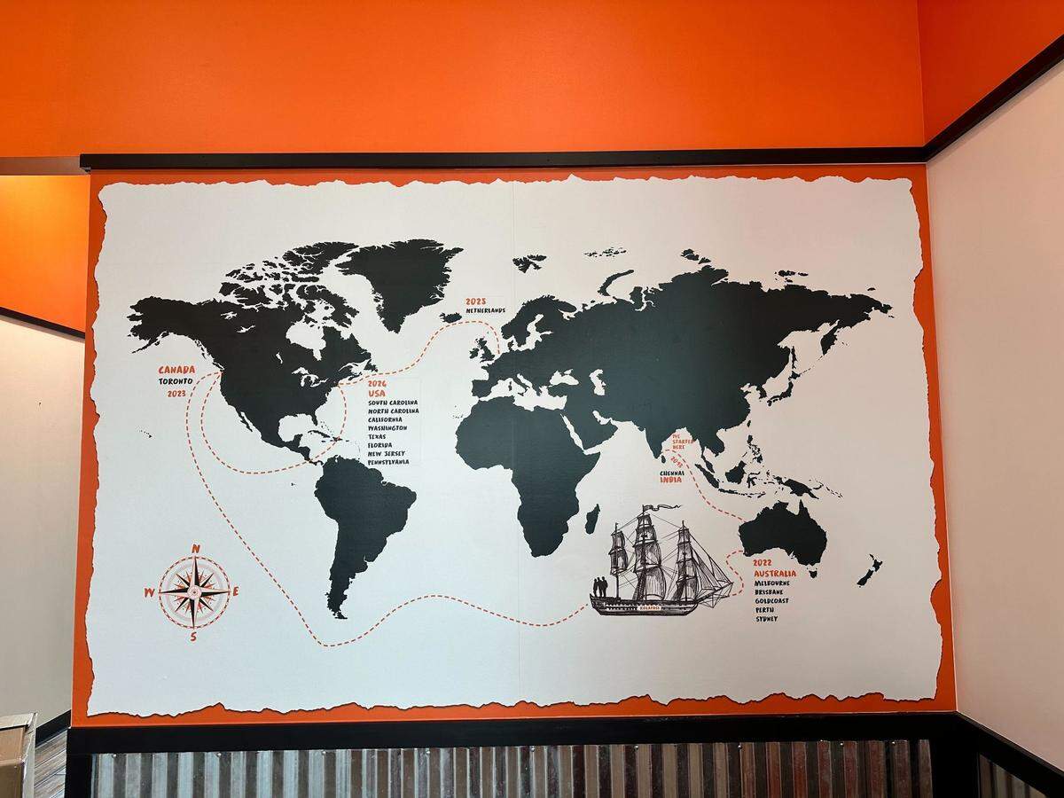 A large mural on a white background with a torn-paper edge depicts a black world map annotated with various international locations and dates. The map includes a red dashed line tracing a path across the oceans, a compass rose, and an illustration of a sailing ship.