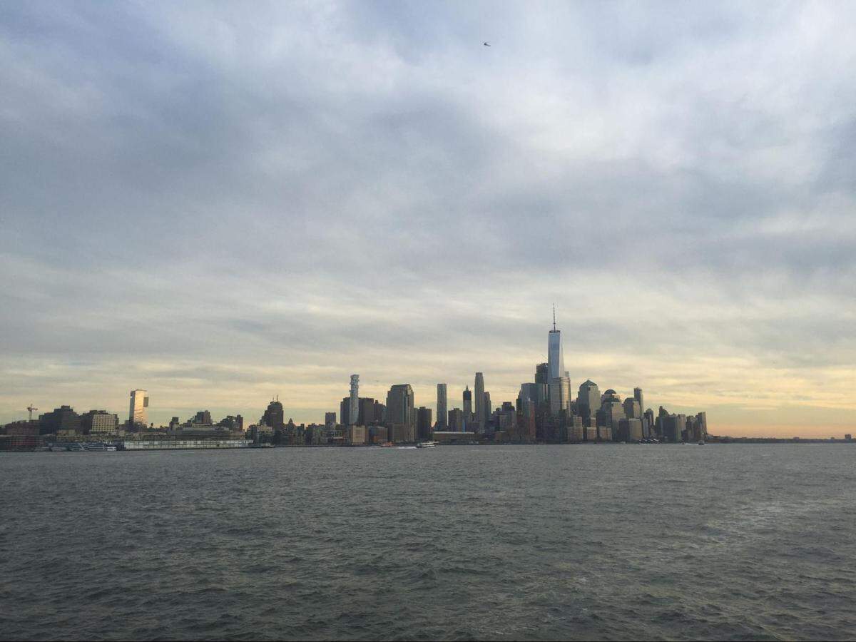 A panoramic, wide-angle shot of the Lower Manhattan skyline as seen from the water. The iconic skyscrapers, including the Freedom Tower, rise against a cloudy, light gray sky with hints of a sunset glow on the horizon. A single bird or plane is visible in the sky. The foreground is dominated by the choppy, dark water of the harbor.