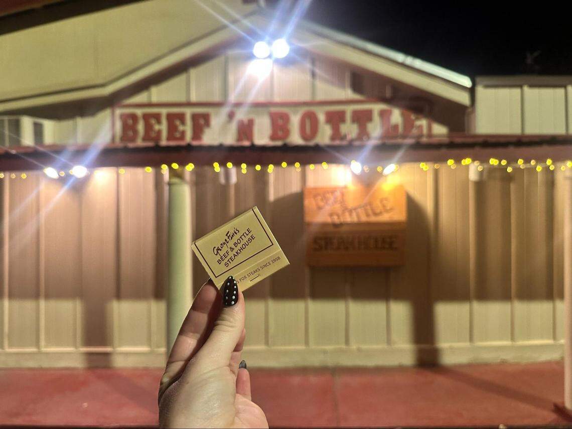 A person’s hand with black and white polka-dot nail art holds up a matchbook for “George & Enid’s Beef & Bottle Steakhouse.” In the background, the warmly lit exterior of the restaurant is visible at night, with a large sign reading “BEEF ‘N BOTTLE” on the building.