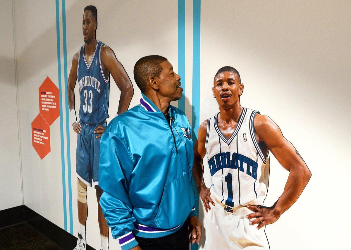 Former Charlotte Hornets player Muggsy Bogues, wearing an original shiny teal team jacket, stands next to a life-sized cutout of his younger self from his playing days, with a cutout of Alonzo Mourning in the background.