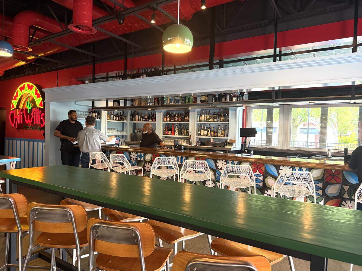 A modern bar area featuring a long wooden counter and white wire-mesh barstools. The base of the bar is decorated with a colorful, geometric flower tile pattern. In the background, a glowing neon “Chili Willis Mexican Cantina” sign hangs on a red wall, and a bartender stands behind a fully stocked back bar.