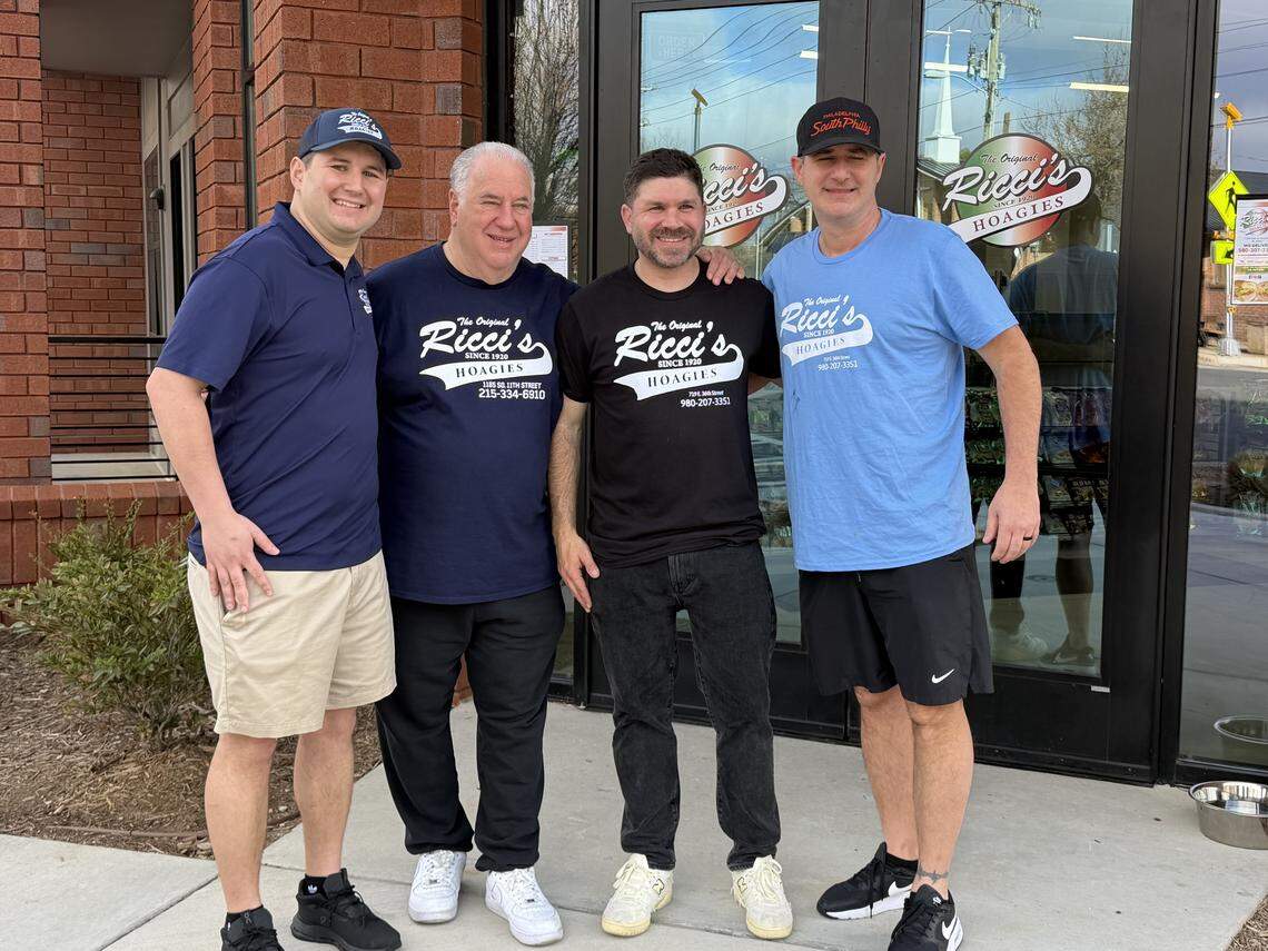Four people are pictured standing in front of a glass entryway branded with “The Original Ricci’s Hoagies”. They are dressed in casual athletic wear, including t-shirts and hats featuring the “Ricci’s Hoagies” logo.