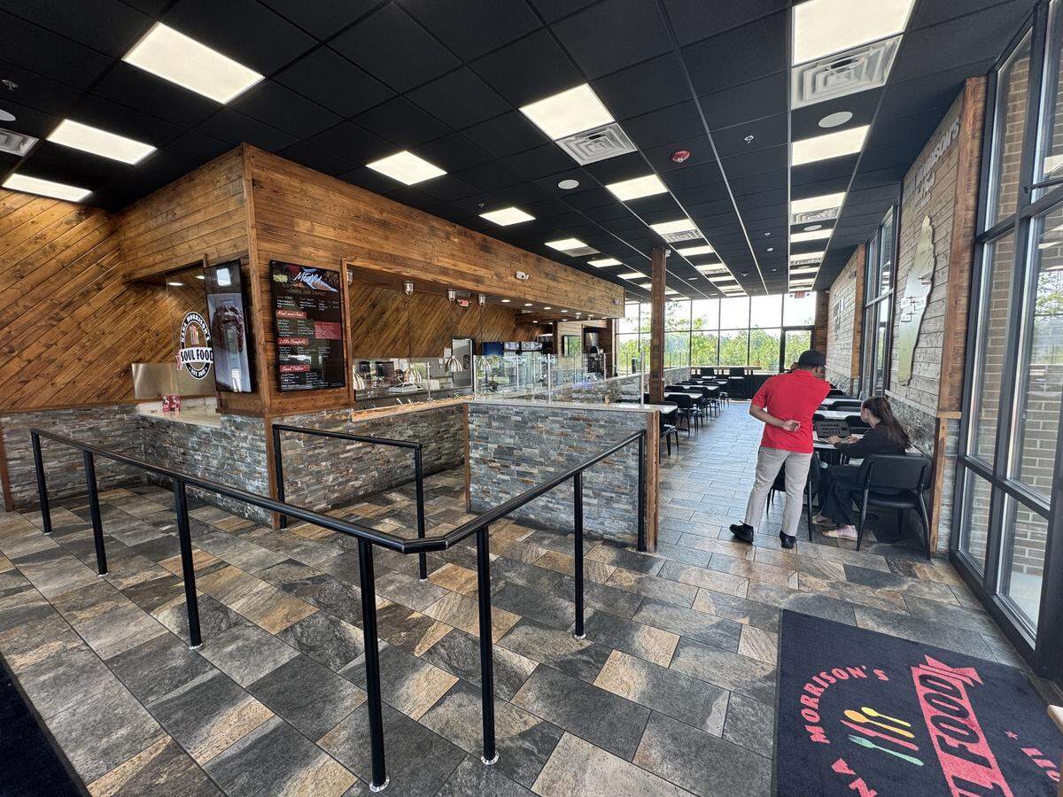 The interior of a spacious, modern restaurant featuring a black grid ceiling with square LED panels and tiled floors. The ordering counter and walls are accented with warm, diagonally oriented wood paneling and a stacked stone facade. Black metal stanchions guide a queue toward the ordering counter on the left. In the dining area to the right, a man in a red polo shirt and khaki pants stands talking to a woman sitting at a table using a laptop. Large floor-to-ceiling windows look out onto greenery in the distance.