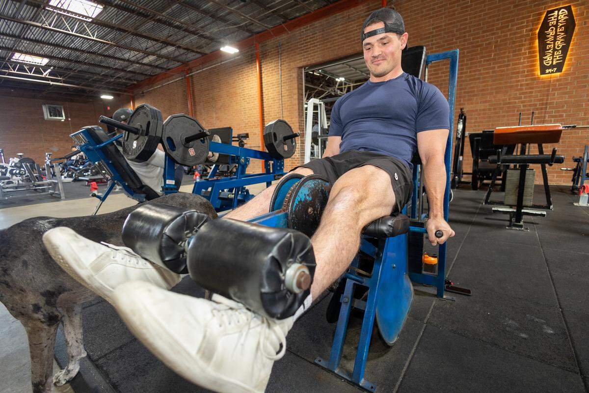 Close-up of a gym user in a navy shirt and cap performing a leg extension on a blue Nautilus machine. A large merle-coated dog’s head and body are partially visible in the foreground.