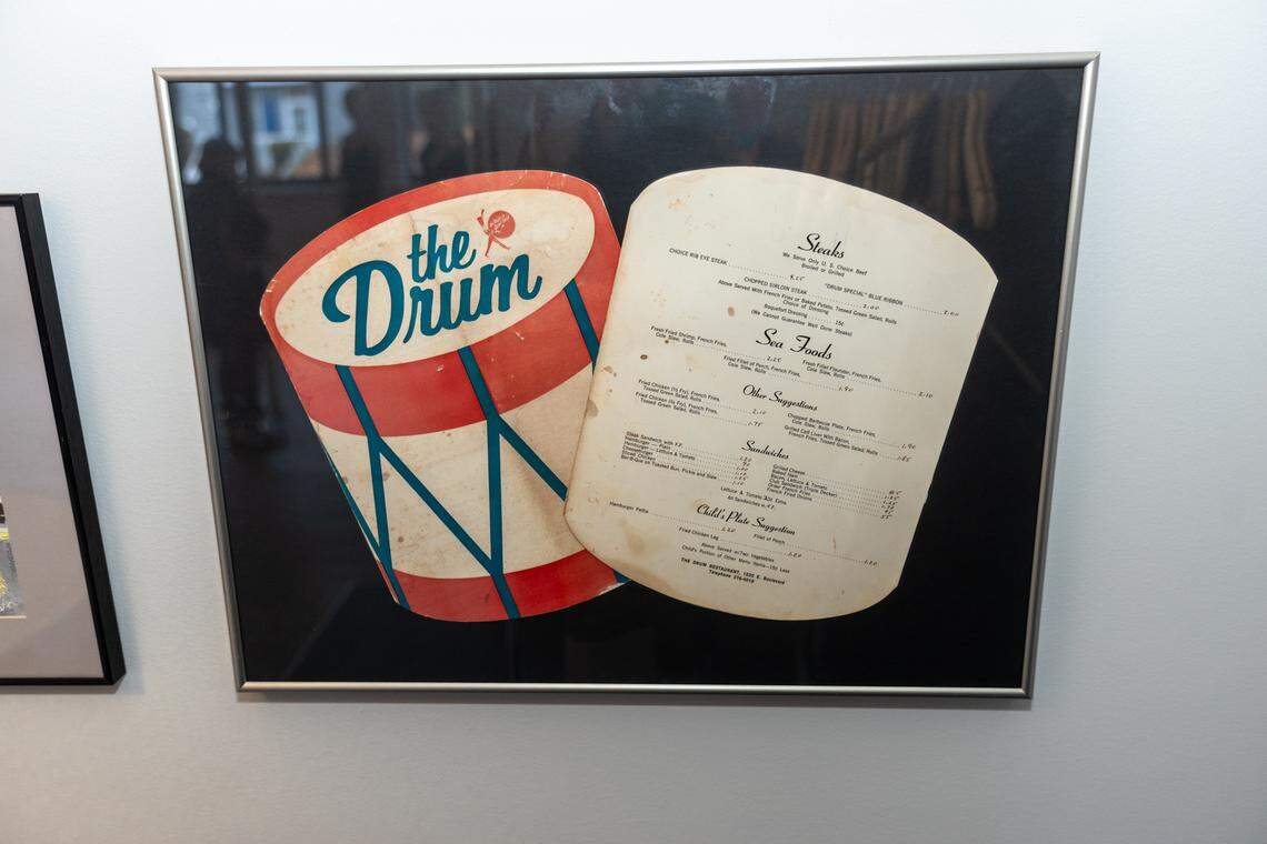 A framed print featuring a vintage, drum-shaped graphic with the text “the Drum” in blue script, next to an old-fashioned paper menu with sections for “Snacks,” “Sea Foods,” “Sandwiches,” and “Chili Plate Suggestions.” The print has a reflection of a person wearing a dark jacket. A portion of another framed artwork depicting a stylized city block is visible on the left.