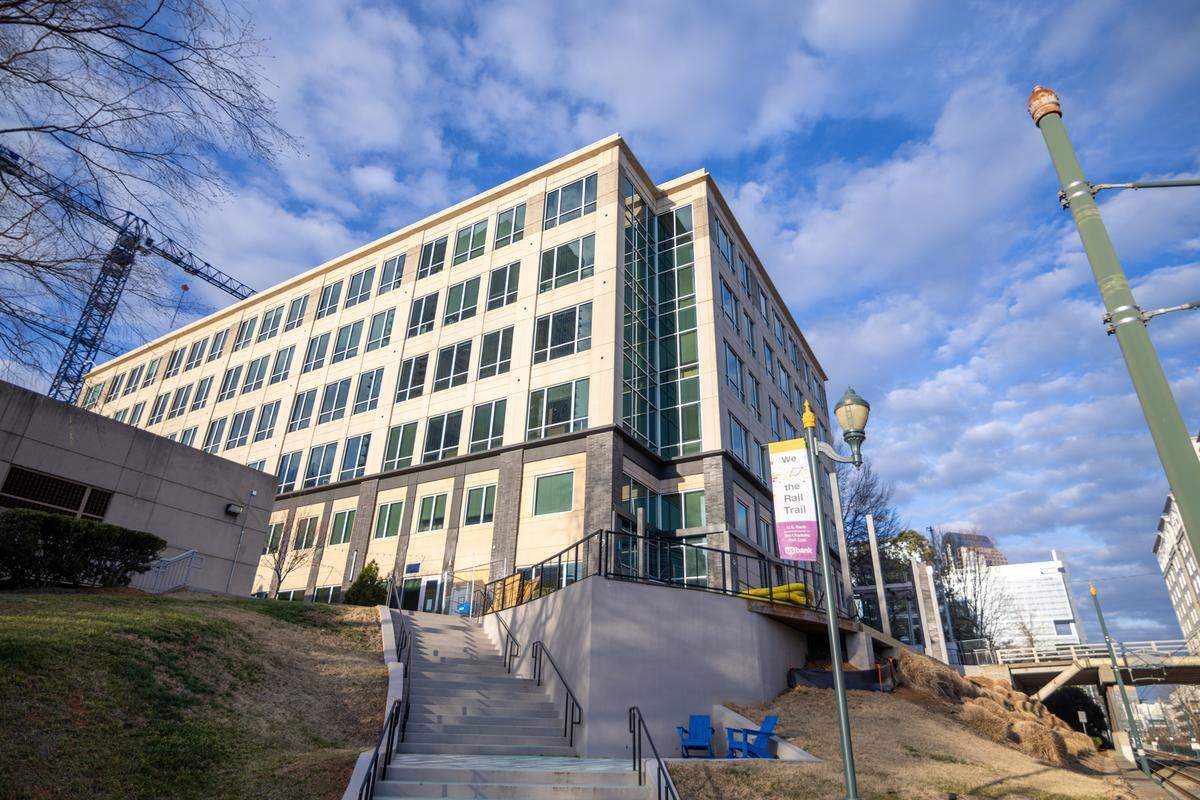 A low-angle exterior shot of a modern, multi-story office or residential building with a tan facade and numerous glass windows. A wide concrete staircase with black metal handrails leads up a grassy hill toward the building’s entrance. In the foreground, a lamppost features a banner that reads “We ❤️ the Rail Trail,” and two blue Adirondack chairs sit on a small landing near the base of the hill. The scene is set under a bright blue sky with wispy white clouds.