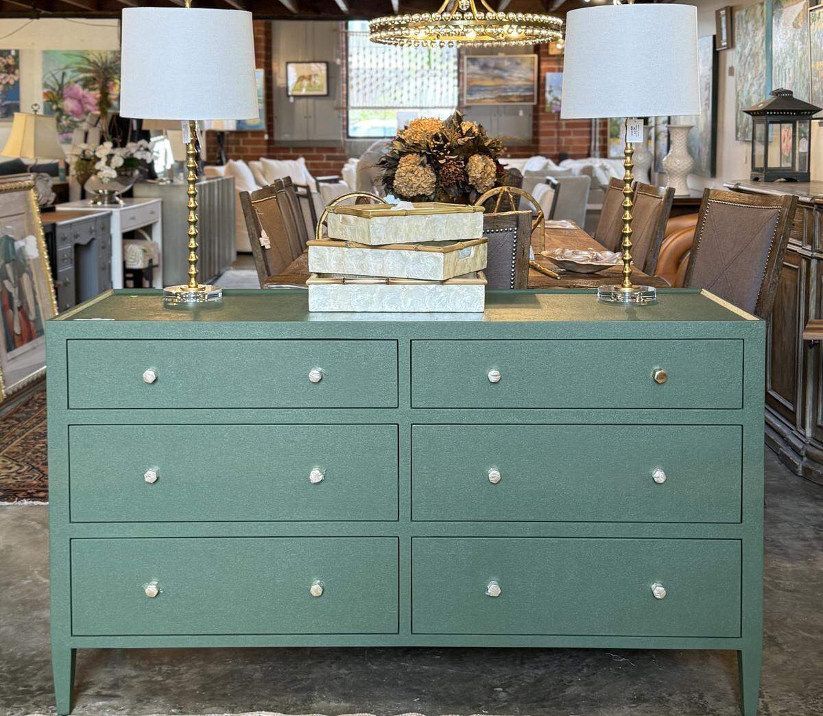 A straight-on view of a wide, six-drawer dresser in a furniture store. The dresser has a modern design with a textured green finish and round silver knobs. On top, a pair of matching lamps with crystal and gold bases flank a centerpiece of stacked boxes and a dried flower arrangement. The background shows the rest of the spacious, loft-style showroom.