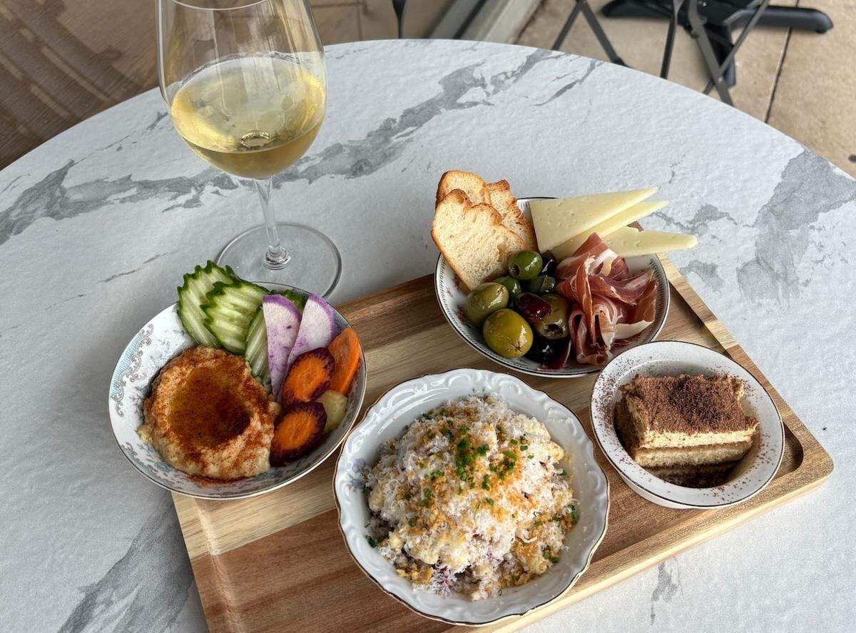 A wooden tray on a white marble table holds a full tapas-style spread and a glass of white wine. The tray includes a bowl of hummus with sliced vegetables, a dish of olives, charcuterie, and cheese, a bowl of creamy pasta topped with shredded cheese and chives, and a small square of tiramisu for dessert.